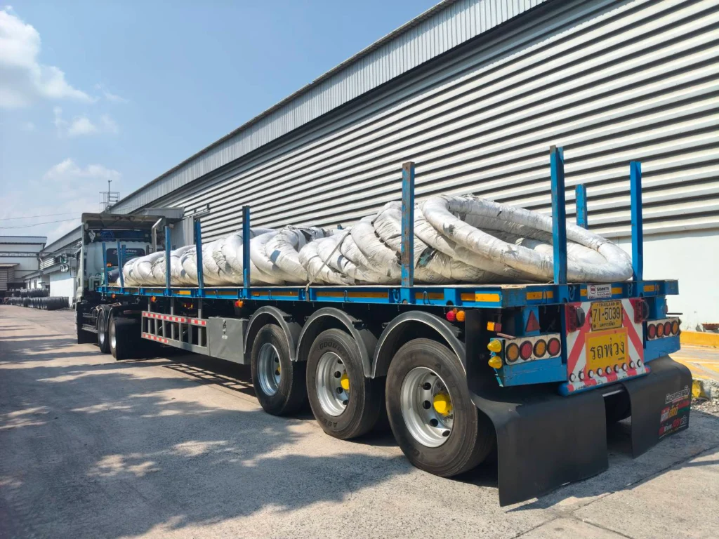 Flatbed truck carrying large white insulated pipes secured with blue stakes beside an industrial building.
