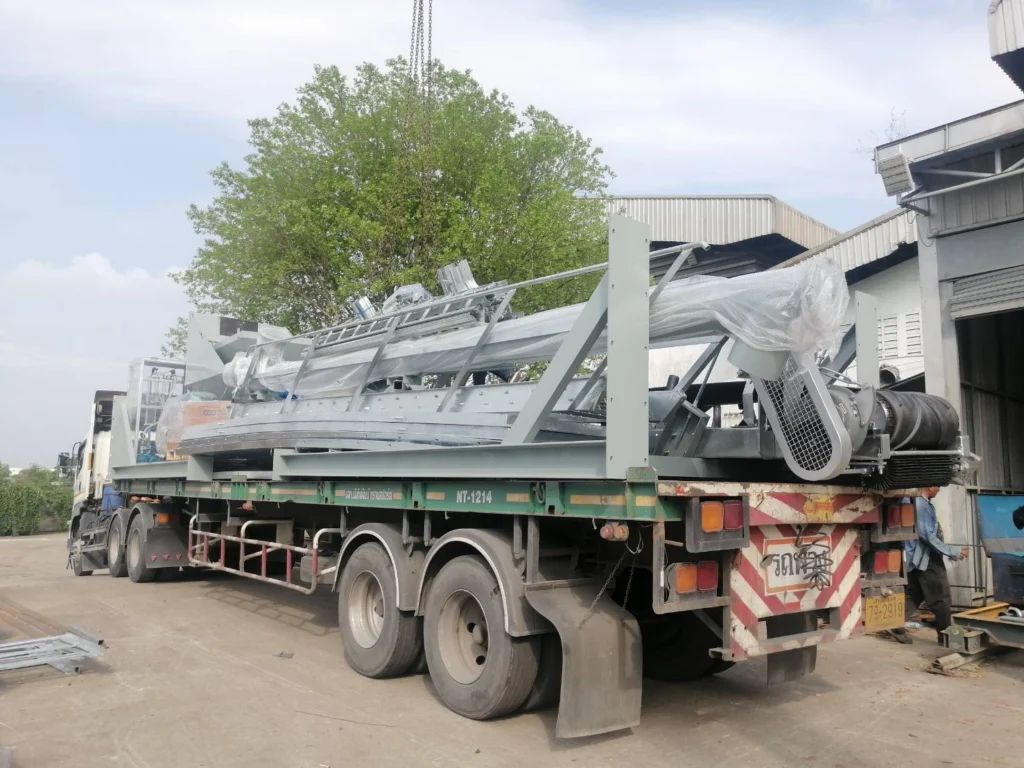 Flatbed truck transporting large bundled steel framework and machinery parts at a loading dock, with wrapped components and ladders secured on top.