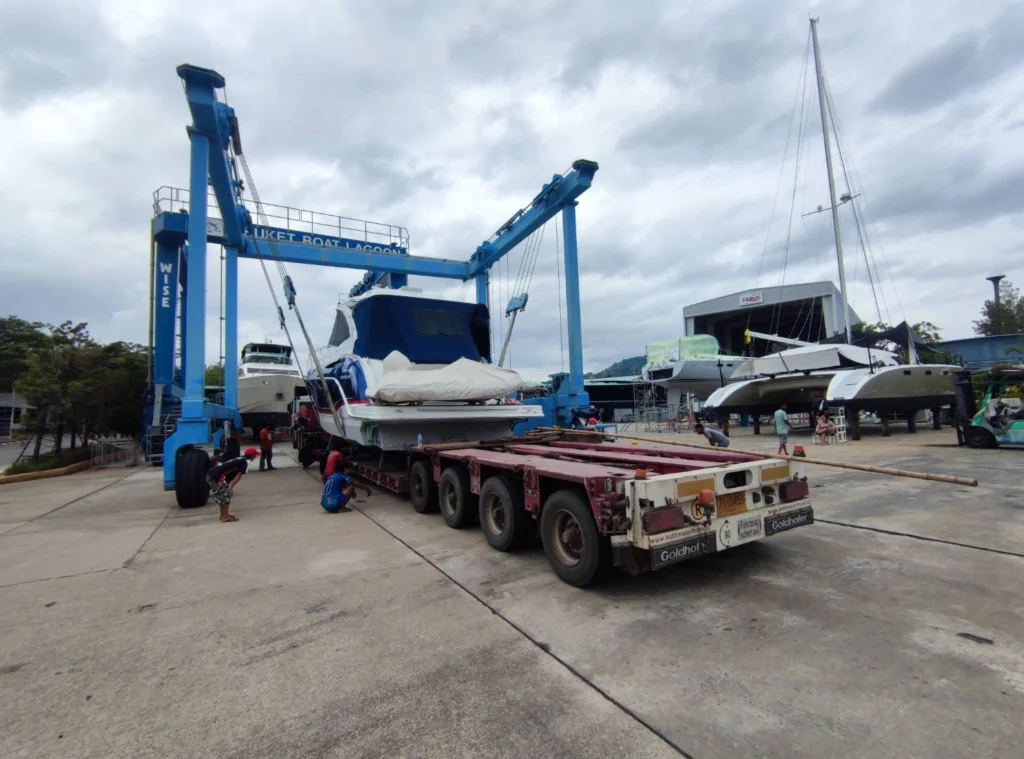 Large blue boat hoist lifts a white-and-blue yacht onto a trailer in a marina repair yard, with workers nearby under a cloudy sky.