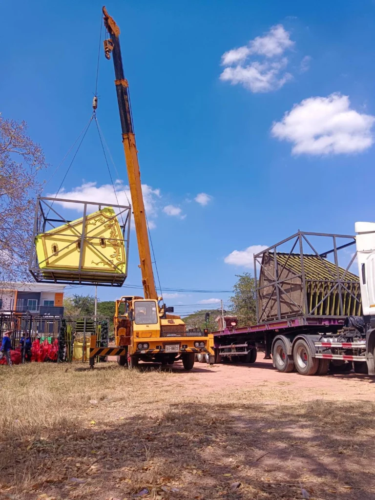 Crane hoisting a large ornate yellow cabinet in a metal crate onto a flatbed truck at an outdoor site on a sunny day.
